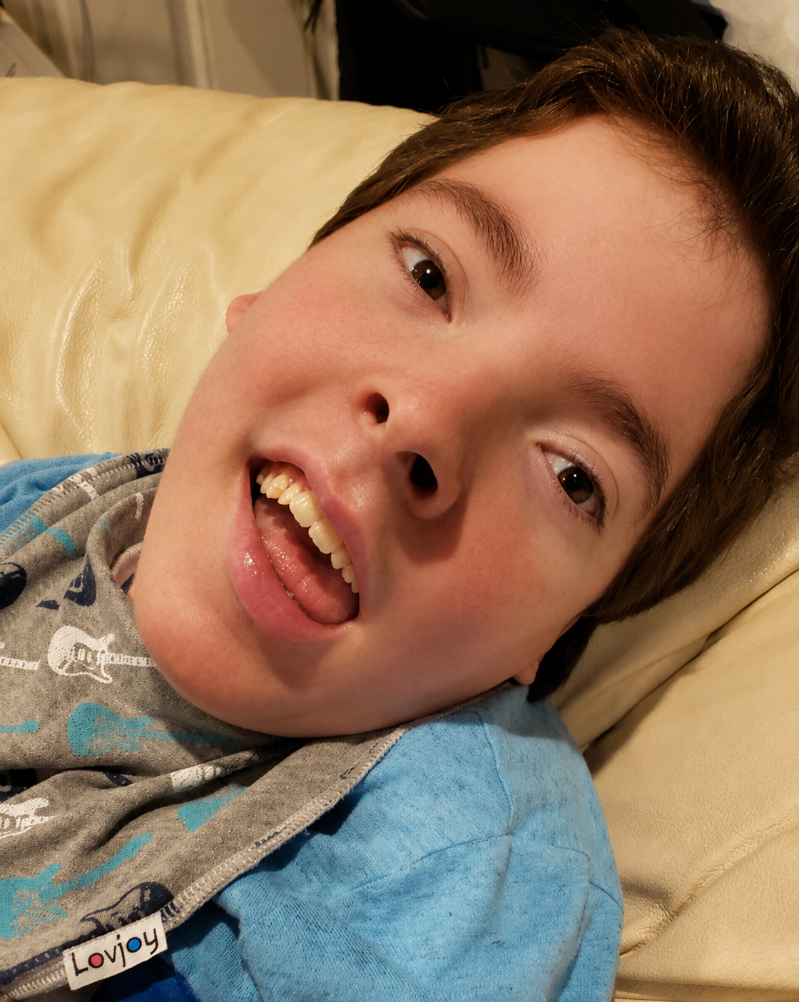 “Greyson Channing smiling joyfully while resting against a cream-colored cushion, wearing a blue shirt.”